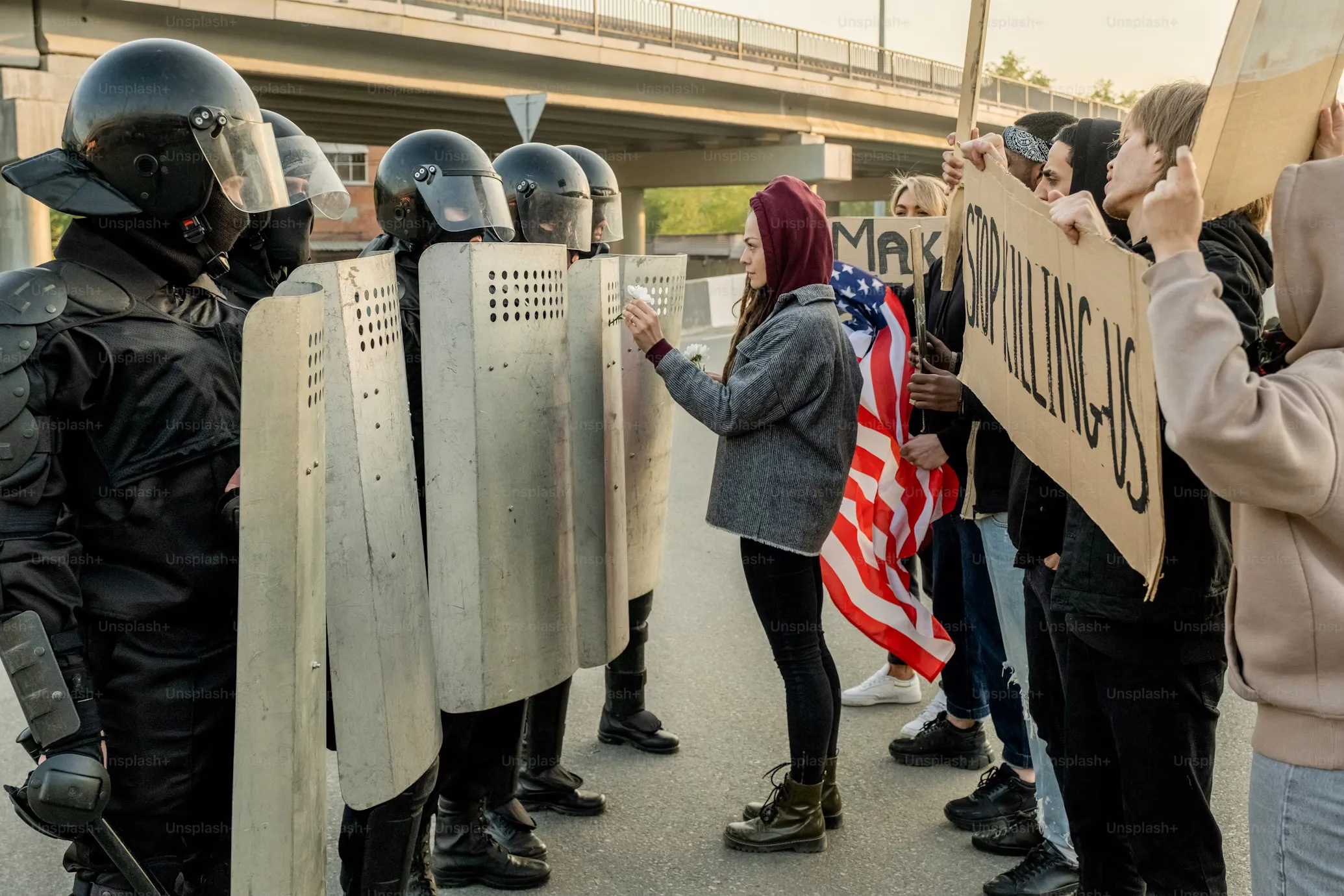 Protester in a head covering faces a line of riot squad law enforcement and places a flower into one of the riot shields