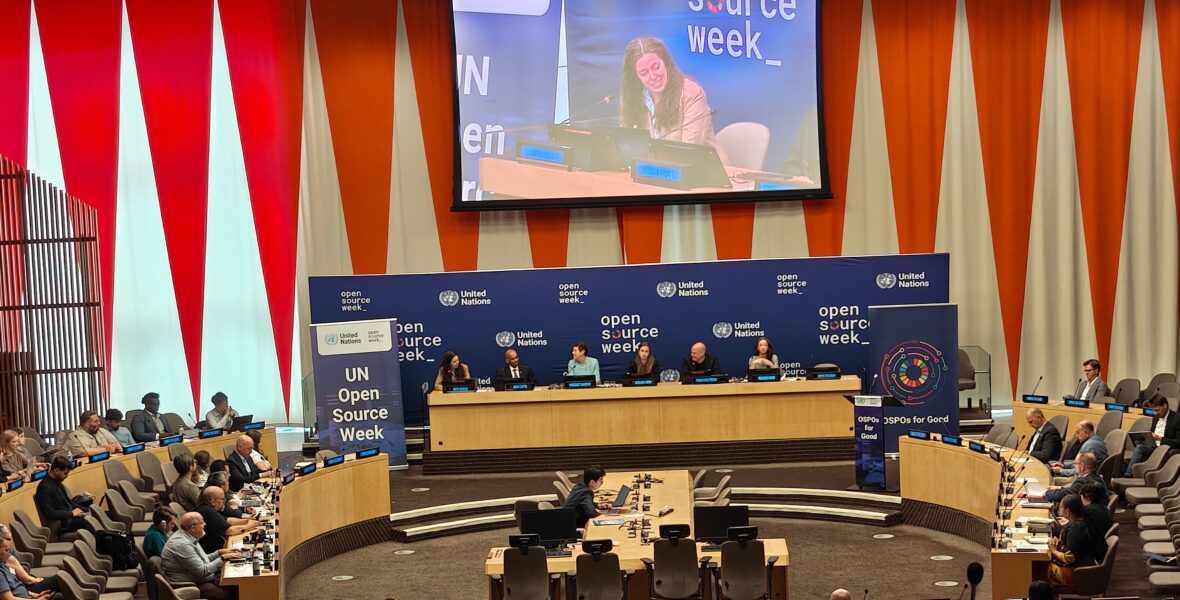 view of a panel discussion at the UN Open Source Week on June 25, 2026. 6 people are seated at the panel with a live feed projected on a screen above them. The audience is seated and flanks the panel to their left and right.