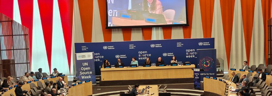 view of a panel discussion at the UN Open Source Week on June 25, 2026. 6 people are seated at the panel with a live feed projected on a screen above them. The audience is seated and flanks the panel to their left and right.